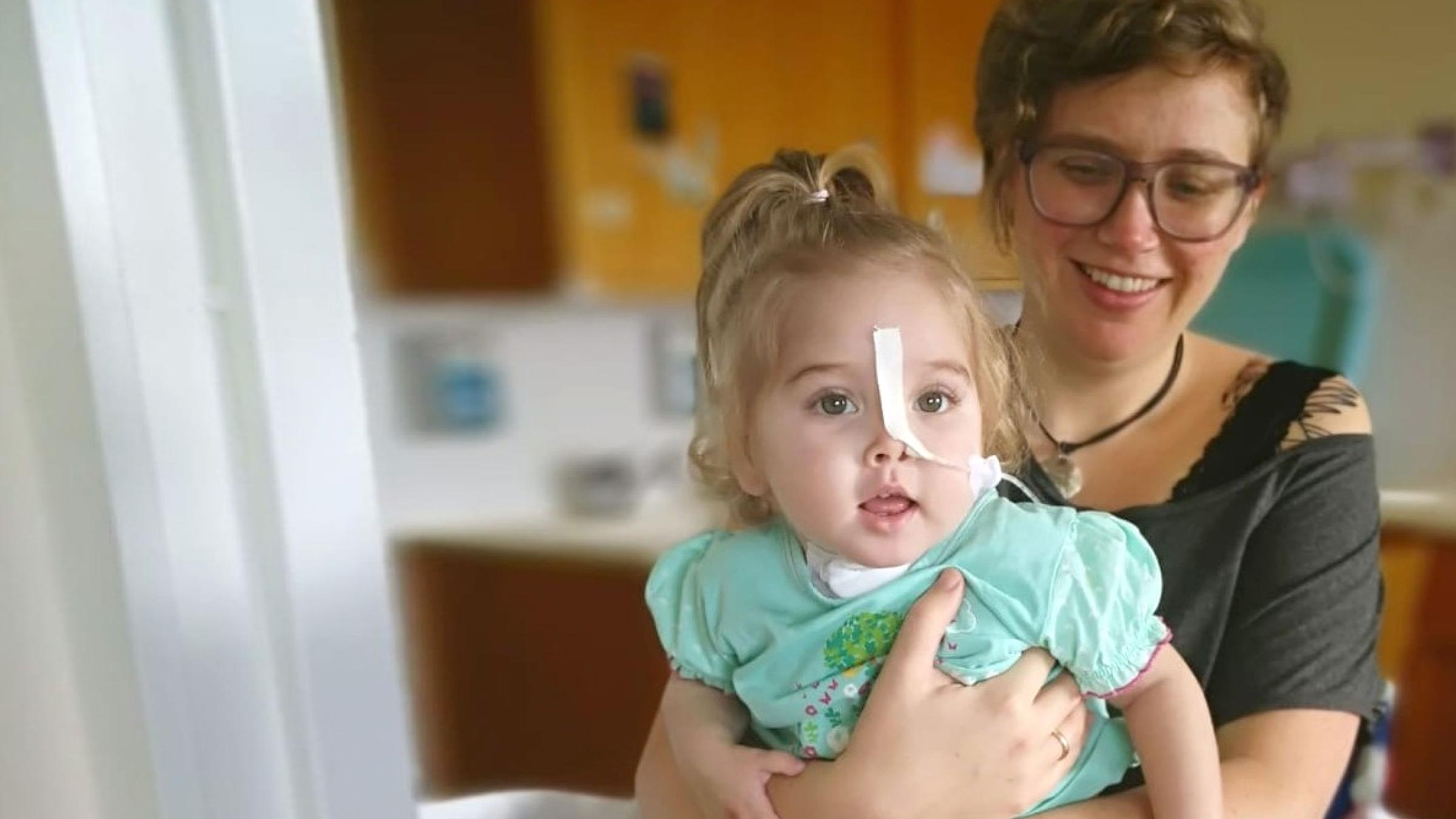 In einem Krankenzimmer hält eine Frau ein Kind in den Armen. Das Kind hat einen Schlauch im Gesicht, der zur Nase führt.