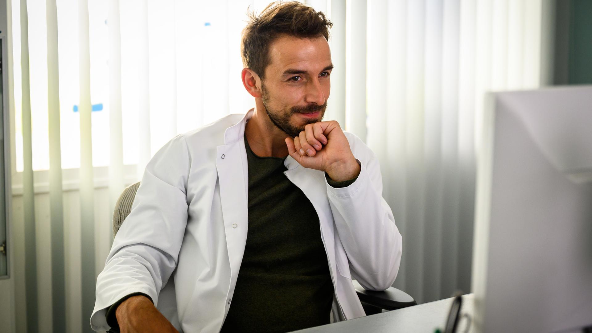 "Bettys Diagnose - Kleine und große Lügen": Dr. Frank Stern (Max Alberti) sitzt an seinem Schreibtisch, den Kopf auf einer Faust abgestützt und schaut zufrieden lächelnd auf seinen Computerbildschirm.