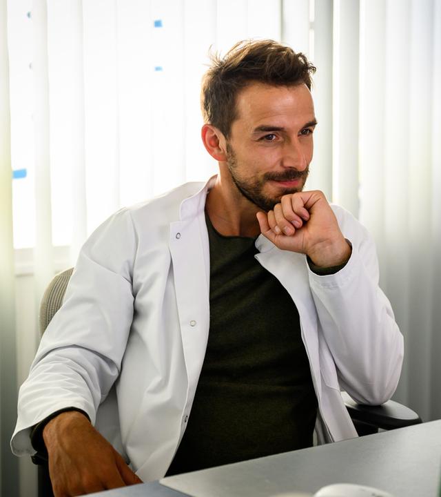 "Bettys Diagnose - Kleine und große Lügen": Dr. Frank Stern (Max Alberti) sitzt an seinem Schreibtisch, den Kopf auf einer Faust abgestützt und schaut zufrieden lächelnd auf seinen Computerbildschirm.