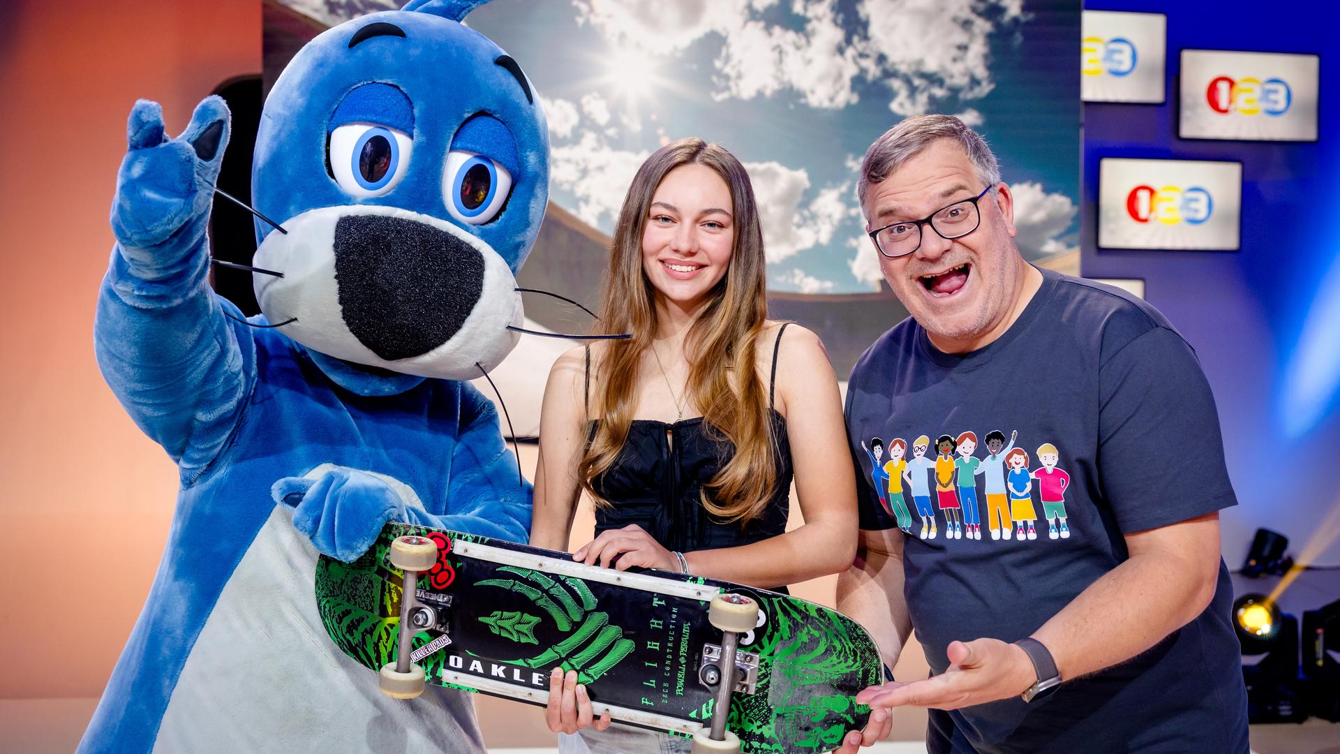 "1, 2 oder 3 - Kleine Helden, große Taten": Skaterin Lilly Stoephasius steht mit ihrem Skateboard in der Hand zwischen Piet Flosse und Elton im Studio.