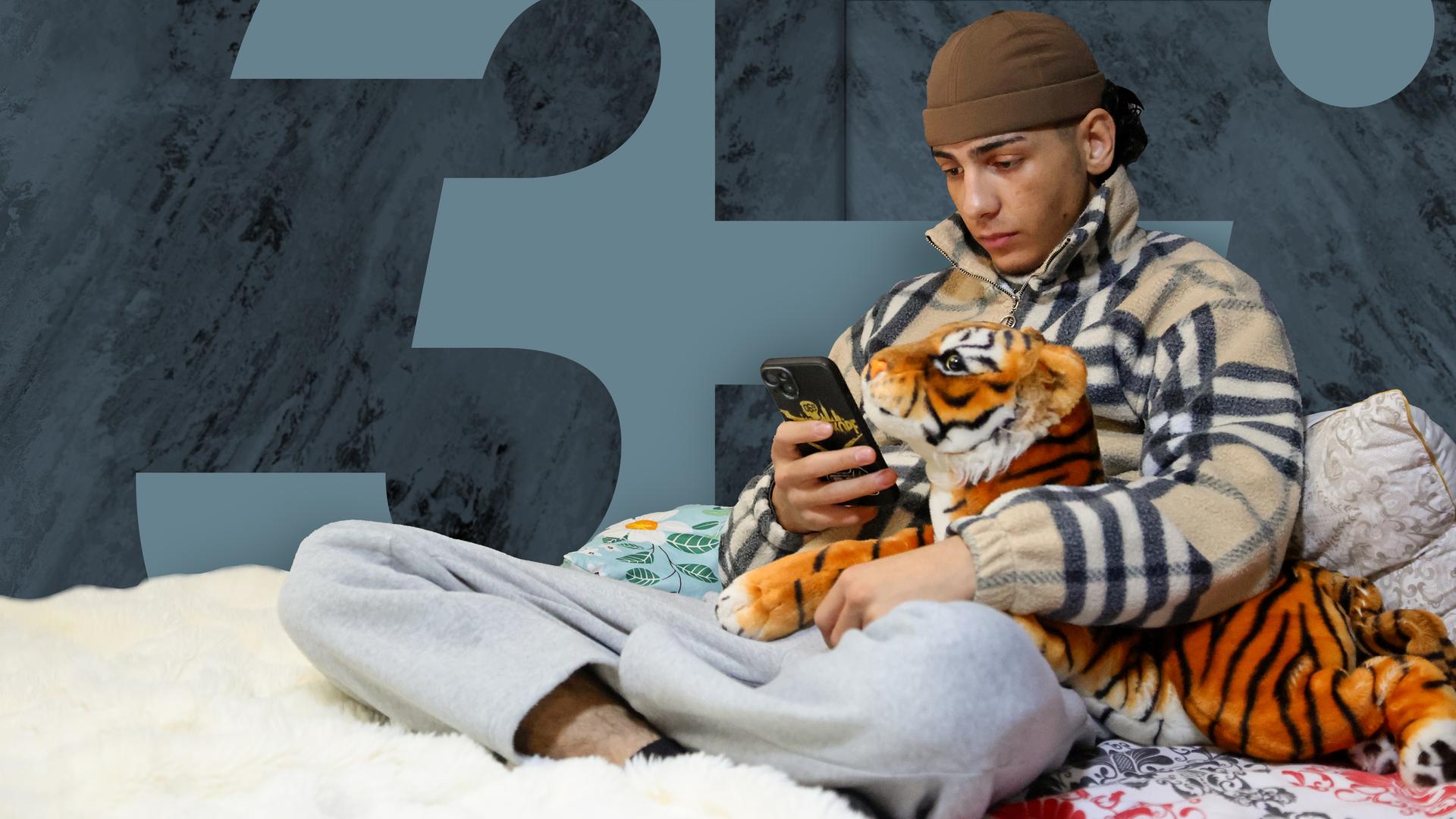 Ein Junge sitzt auf einem Bett und schaut auf sein Handy. In einer Hand hält er das Smartphone, in der anderen ein Kuscheltier.