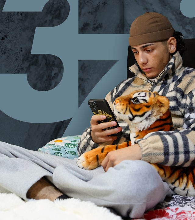 Ein Junge sitzt auf einem Bett und schaut auf sein Handy. In einer Hand hält er das Smartphone, in der anderen ein Kuscheltier.