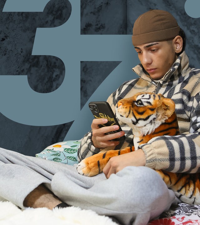 Ein Junge sitzt auf einem Bett und schaut auf sein Handy. In einer Hand hält er das Smartphone, in der anderen ein Kuscheltier.