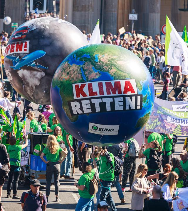 Auf einer Demonstration für die Rettung des Klimas wuseln viele Menschen mit Flaggen, Bannern und zwei riesigen Luftballons in Form der Erde durcheinander.