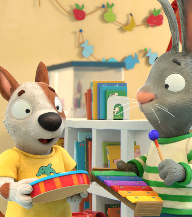 Zac steht links mit seiner orange-rot-gestreiften Trommel, in der Mitte Pip mit einem bunten Xylophon und rechts Posy mit einer Rassel. Die drei unterhalten sich. Im Hintergrund des Kinderzimmers ist ein Regal mit Büchern zu sehen und zwei Girlanden an der Wand.