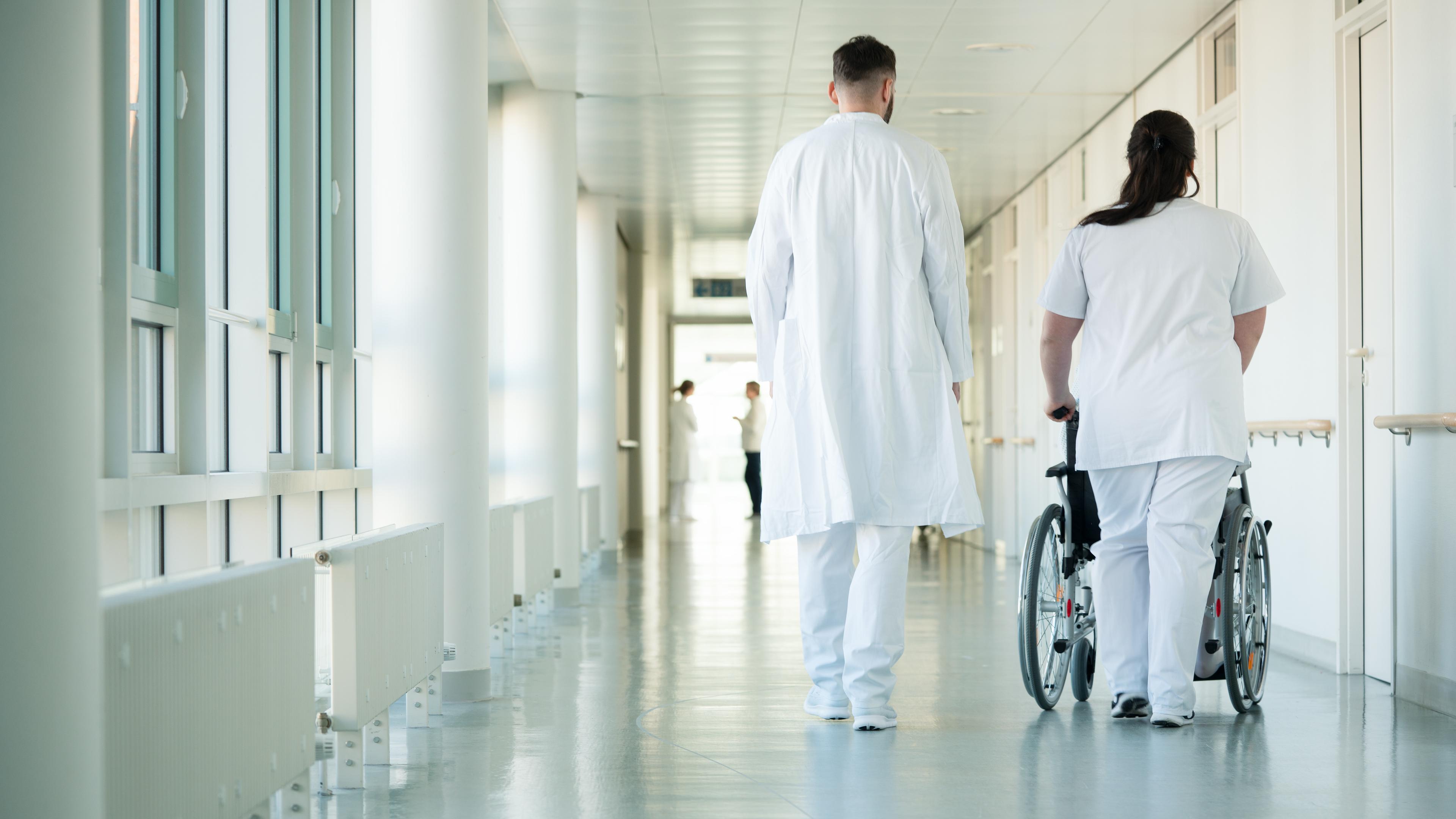 Arzt und Pflegerin, die einen Rollstuhl schiebt, gehen den Gang eines Krankenhauses hinunter.