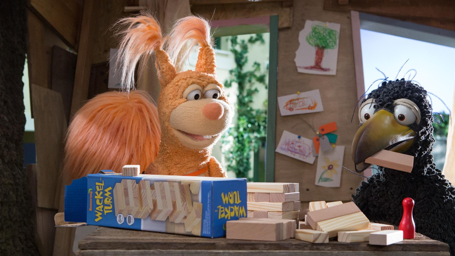 Rabe Rudi und Eichhörnchen Ella sitzen im Baumhaus, Rudi hat einen Klotz im Schnabel, weitere Klötze samt Spielverpackung liegen vor ihnen.