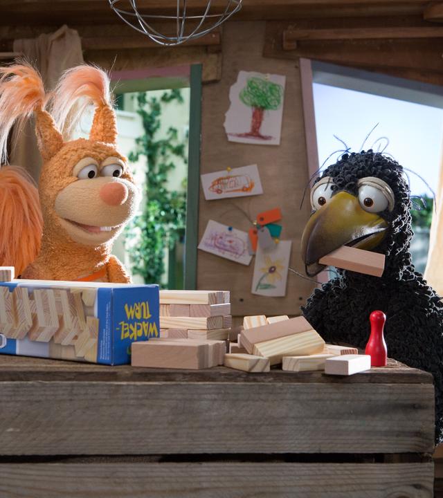 Rabe Rudi und Eichhörnchen Ella sitzen im Baumhaus, Rudi hat einen Klotz im Schnabel, weitere Klötze samt Spielverpackung liegen vor ihnen.