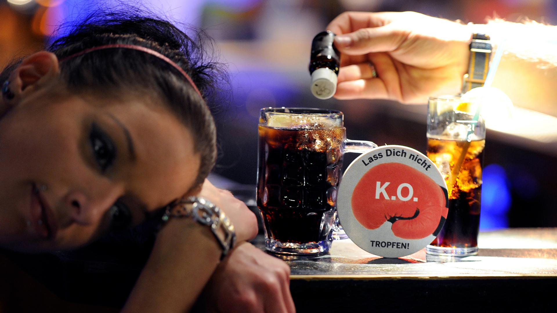 Eine Frau hat den Kopf auf den Tresen gelegt, während neben ihr eine Hand ein kleines braunes Fläschchen über ihr Glas hält.