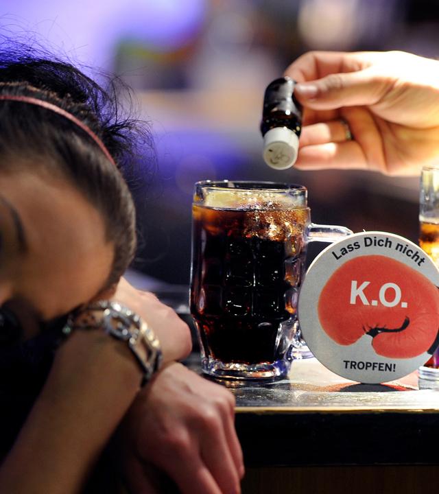 Eine Frau hat den Kopf auf den Tresen gelegt, während neben ihr eine Hand ein kleines braunes Fläschchen über ihr Glas hält.