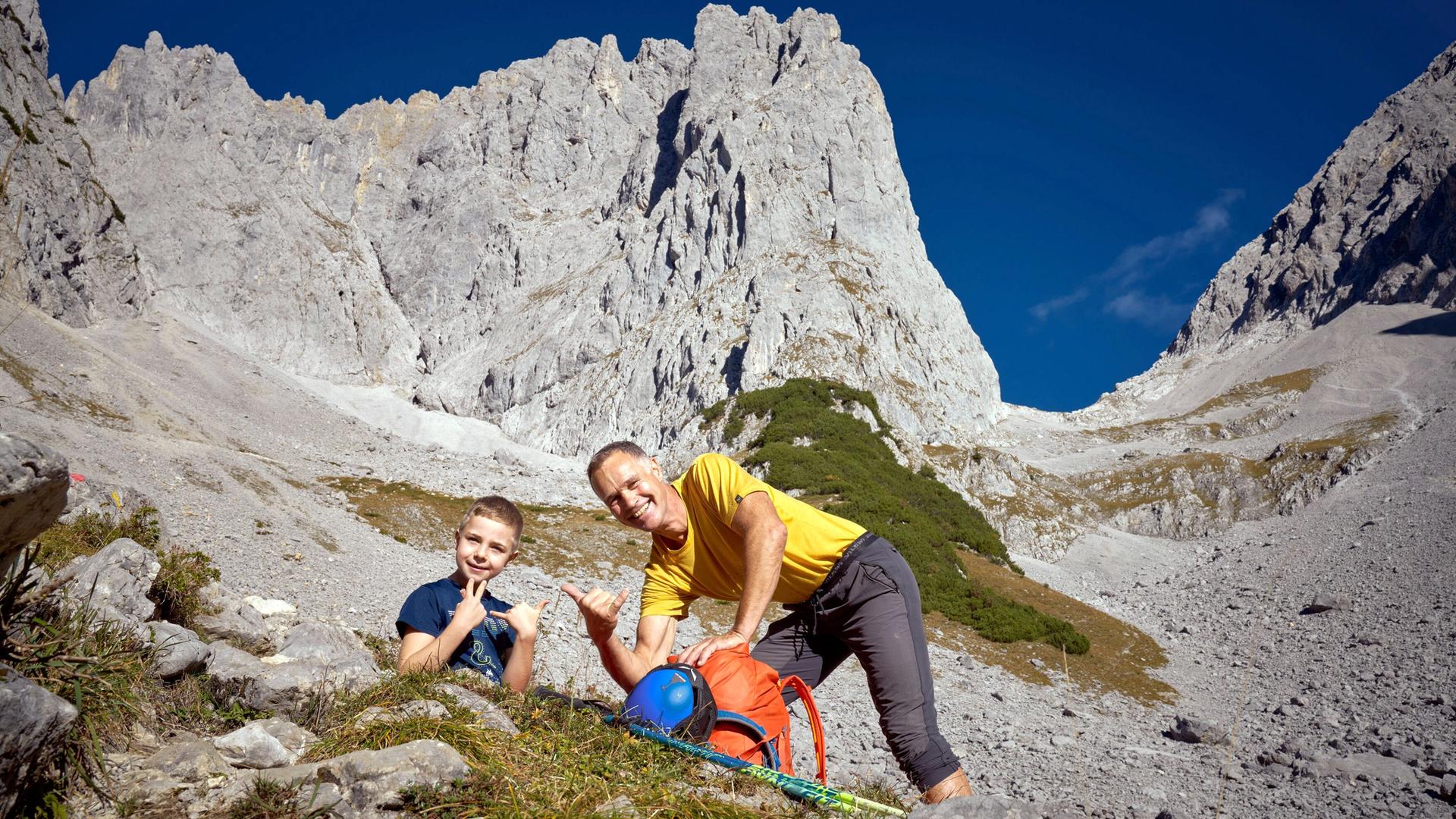 Das Bild zeigt zwei Personen in einer alpinen Landschaft. Im Vordergrund sitzen ein älterer Mann und ein Junge auf einem steinigen Untergrund, umgeben von Gräsern und niedrigen Pflanzen. Der Mann trägt ein gelbes T-Shirt und graue Hose, während der Junge ein blaues T-Shirt trägt. Beide posieren freundlich, der Junge zeigt mit beiden Händen ein Zeichen des "Shaka", während der Mann mit einer Hand das gleiche Zeichen macht.  Im Hintergrund erhebt sich eine massive Felsformation, die aus grauem Gestein besteht und steil aufsteigt. Der Himmel ist klar und blau, und im unteren Drittel des Bildes sind einige flachere, grüne Bereiche sichtbar, die mit Bäumen bewachsen sind. Das Gesamtbild vermittelt einen Eindruck von Weite und der Schönheit der alpinen Natur, während es gleichzeitig eine Elemente des Bergsteigens und der Outdoor-Aktivitäten zeigt.