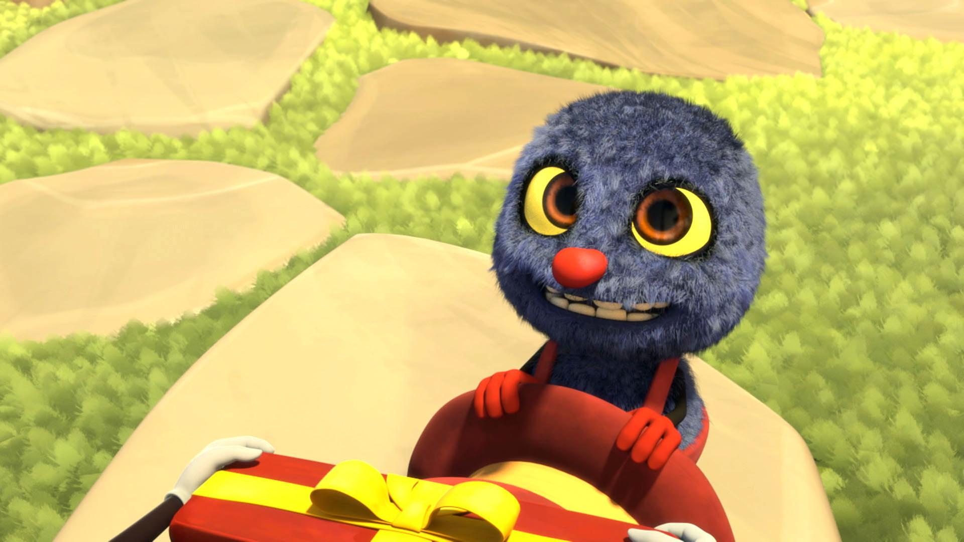 Klaus hat seinen Hut ausgezogen und hält ihn in den Händen. Im unteren Bildrand sieht man ein längliches Geschenk, das ihm überreicht wird. Er wirkt glücklich und gespannt.