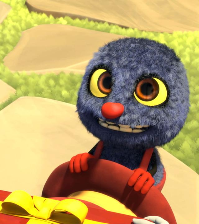 Klaus hat seinen Hut ausgezogen und hält ihn in den Händen. Im unteren Bildrand sieht man ein längliches Geschenk, das ihm überreicht wird. Er wirkt glücklich und gespannt.