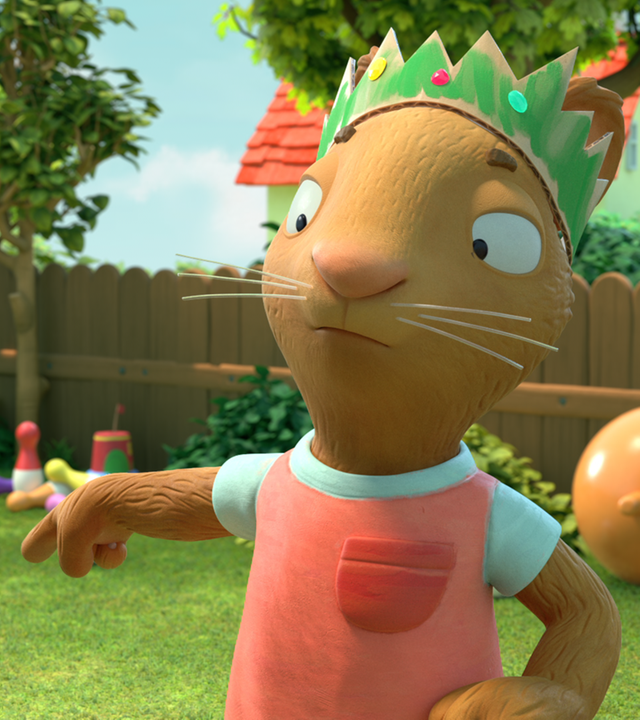 Posy, mit bunter Krone auf dem Kopf, streckt ihren rechten Arm aus und zeigt im Garten auf auf dem Boden liegende Kegel. Rechts im Hintergrund liegt ein Hüpfball.