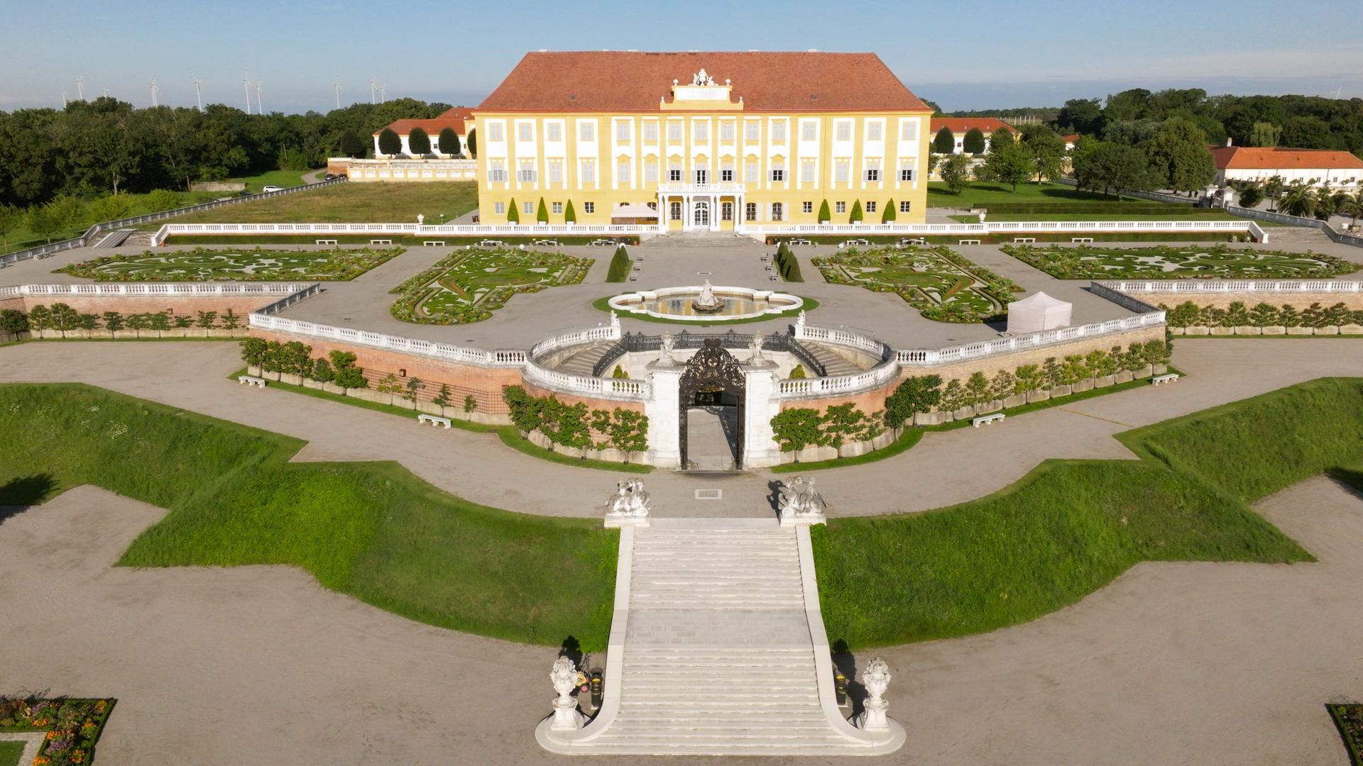 Das Bild zeigt eine weitläufige Gartenlandschaft mit einem großen, gelben Schloss im Hintergrund. Das Schloss hat eine symmetrische Fassade mit vielen Fenstern. Vor dem Schloss befindet sich ein großzügiger Garten mit geometrischen Beeten, die sorgsam angelegt sind. In der Mitte des Gartens ist ein runder Brunnen mit Skulpturen, umgeben von einer Mauer mit Pflanzen. Breite, steinerne Stufen führen von der Gartenterrasse hinunter zum unteren Bereich des Gartens. Die Umgebung besteht aus gepflegtem Grün mit vereinzelten Bäumen und Büschen und erzeugt eine ruhige, einladende Atmosphäre. Der Himmel ist klar und sonnig.