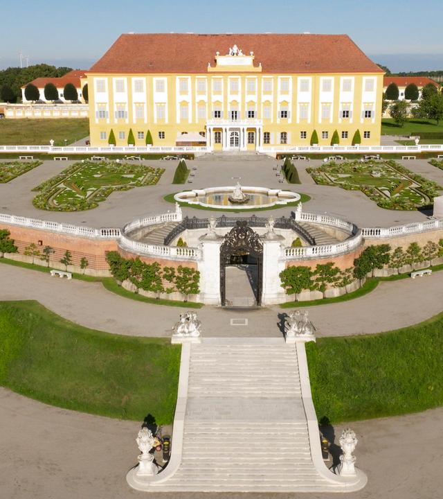 Das Bild zeigt eine weitläufige Gartenlandschaft mit einem großen, gelben Schloss im Hintergrund. Das Schloss hat eine symmetrische Fassade mit vielen Fenstern. Vor dem Schloss befindet sich ein großzügiger Garten mit geometrischen Beeten, die sorgsam angelegt sind. In der Mitte des Gartens ist ein runder Brunnen mit Skulpturen, umgeben von einer Mauer mit Pflanzen. Breite, steinerne Stufen führen von der Gartenterrasse hinunter zum unteren Bereich des Gartens. Die Umgebung besteht aus gepflegtem Grün mit vereinzelten Bäumen und Büschen und erzeugt eine ruhige, einladende Atmosphäre. Der Himmel ist klar und sonnig.