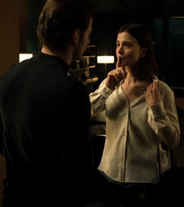 "Serial Cleaner - Kollisionskurs": In einem dunklen Innenraum stehen sich Teri Marquard (Aylin Tezel) und Vincent Karanz (Stefan Gorski) gegenüber, Tischleuchten im Hintergrund spenden spärliches Licht. Teri hält ihren Zeigefinger an ihren Mund, damit Vincent keinen Ton von sich gibt. Sie zeigt Vincent, dass sie auf der linken Brustseite verkabelt ist.