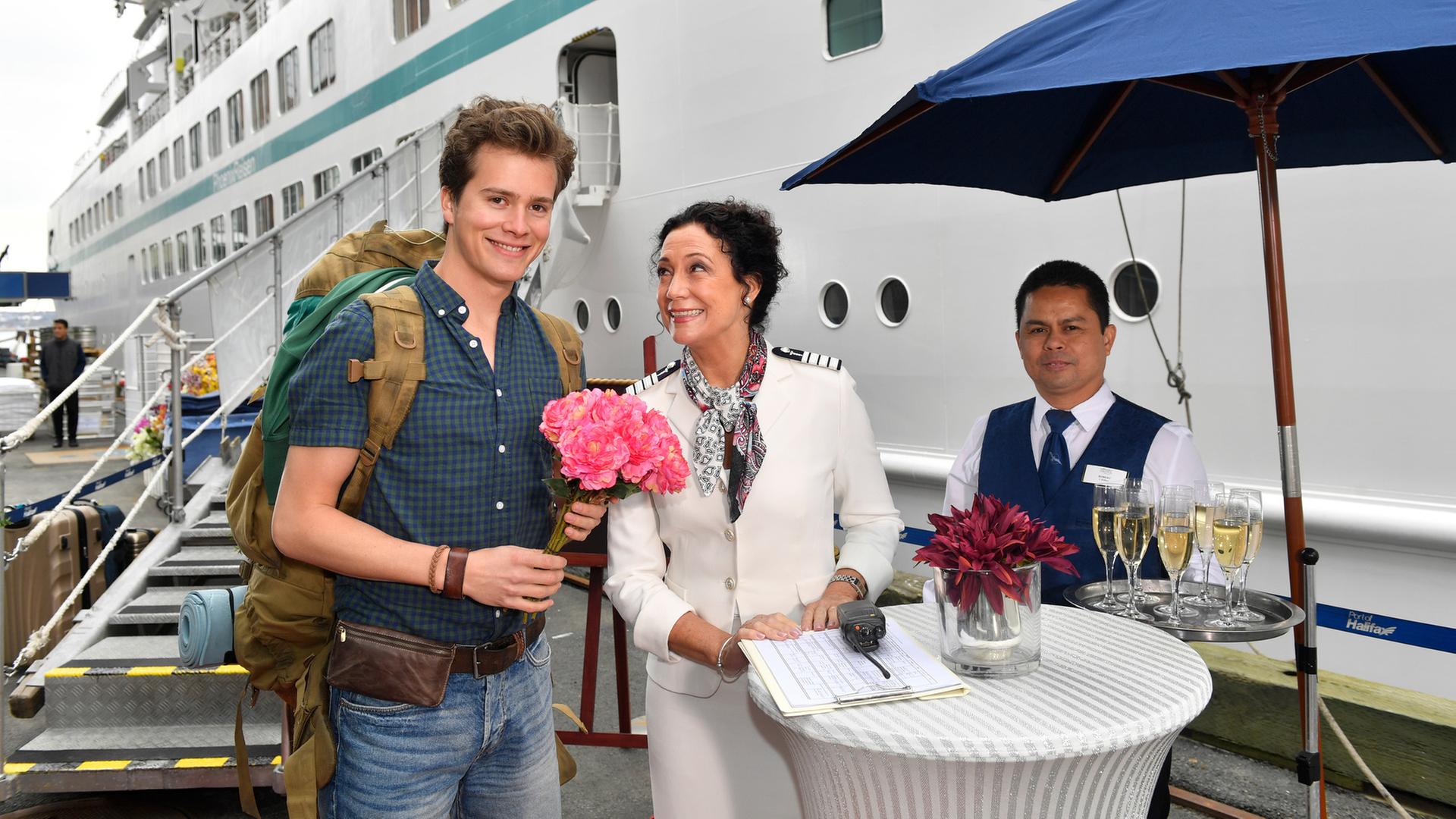 "Das Traumschiff: Kolumbien": Eric Sommer (Lucas Bauer) hält Hanna Liebhold (Barbara Wussow) einen Bund pinke Blumen entgegen, die ihn freundlich anlächelt. Neben ihr steht ein weiterer Bediensteter des Schiffs (Darsteller unbekannt), der ein Tablett mit Sektgläsern hält.