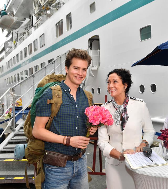 "Das Traumschiff: Kolumbien": Eric Sommer (Lucas Bauer) hält Hanna Liebhold (Barbara Wussow) einen Bund pinke Blumen entgegen, die ihn freundlich anlächelt. Neben ihr steht ein weiterer Bediensteter des Schiffs (Darsteller unbekannt), der ein Tablett mit Sektgläsern hält.