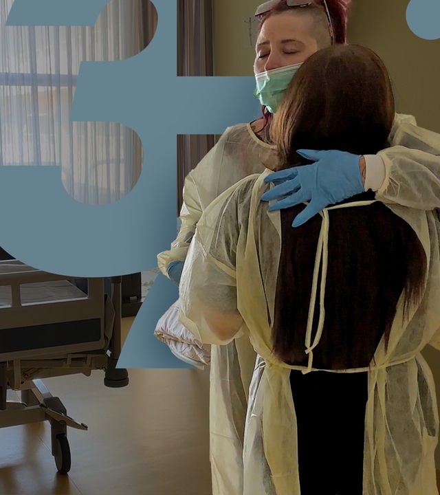 Im Bett eines Krankenzimmers liegt Doris. Ihre Tochter und ihre Enkelin stehen daneben, tragen Schutzkleidung und umarmen sich.