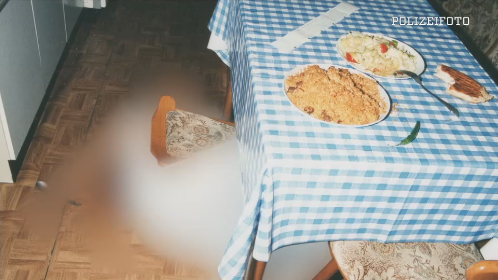 Tatortfoto: Leiche (unkenntlich) liegt am Boden einer Küche. Auf dem Tisch stehen zwei Teller mit Essen und ein angebissenes Stück Brot. Oben Rechts eingeblendet: "Polizeifoto"
