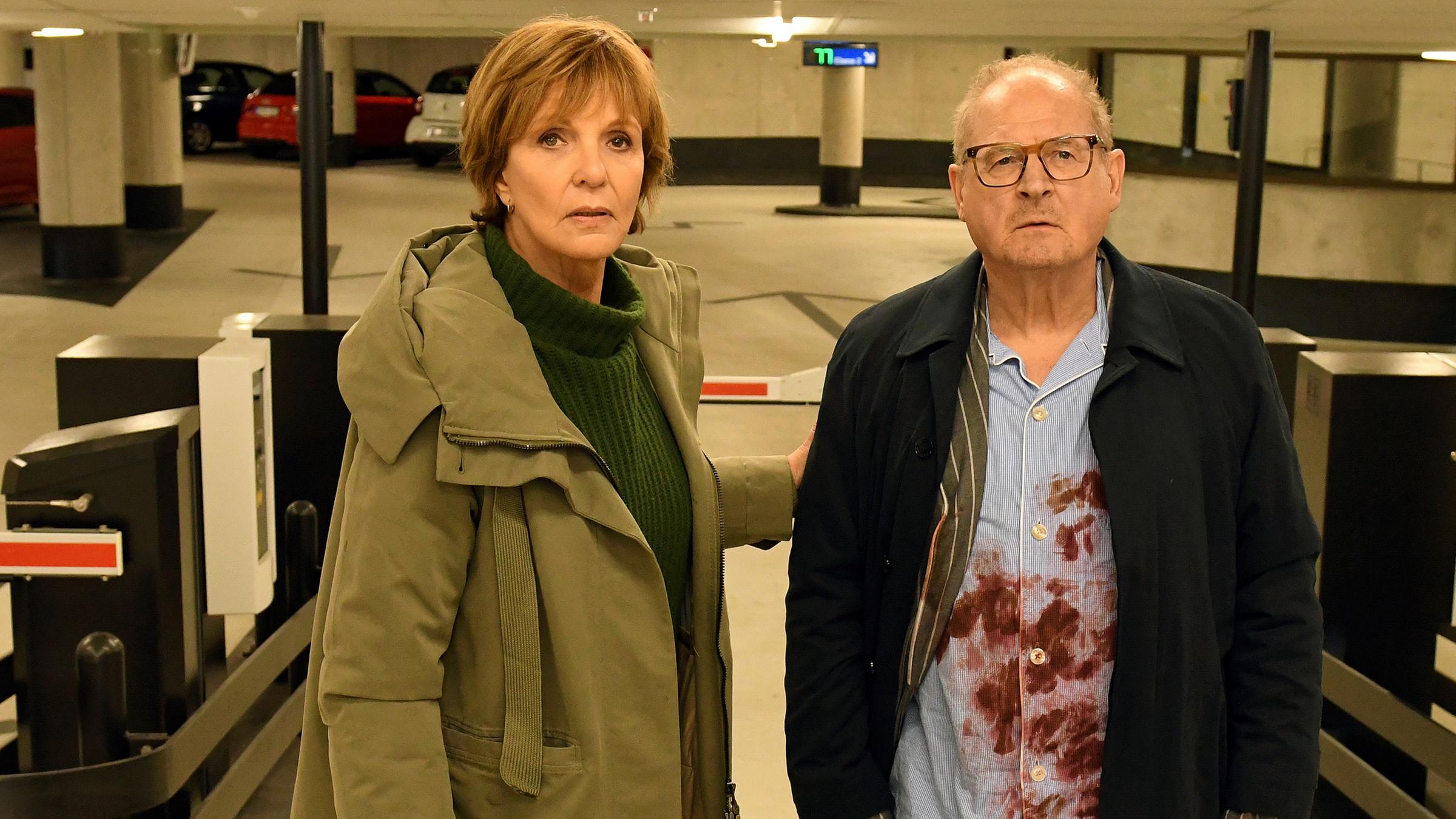 Ellen Lucas (Ulrike Kriener) und Wolfgang Löhns (Burghart Klaußner) stehen nebeneinander in einem Parkhaus vor der Einfahrtsschranke. Ellen Lucas hat die Hand auf Wolfgang Löhns' Arm gelegt, der unter seinem Mantel einen blutverschmierten Schlafanzug trägt.