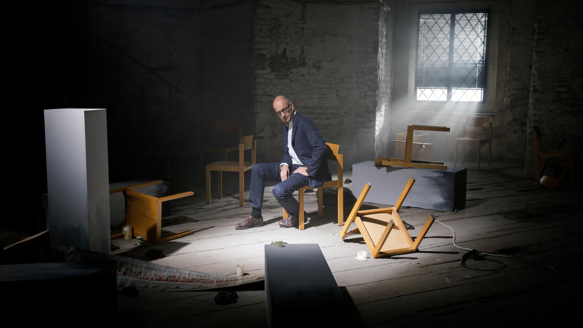 Ein Mann in Anzug sitzt nachdenklich auf einem Stuhl in einem dunklen Raum mit umgestürzten Möbeln und diffusem Licht. Eine Lichtquelle fällt durch ein vergittertes Fenster auf ihn.