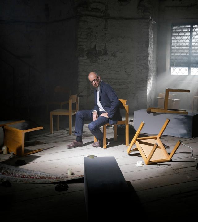 Ein Mann in Anzug sitzt nachdenklich auf einem Stuhl in einem dunklen Raum mit umgestürzten Möbeln und diffusem Licht. Eine Lichtquelle fällt durch ein vergittertes Fenster auf ihn.