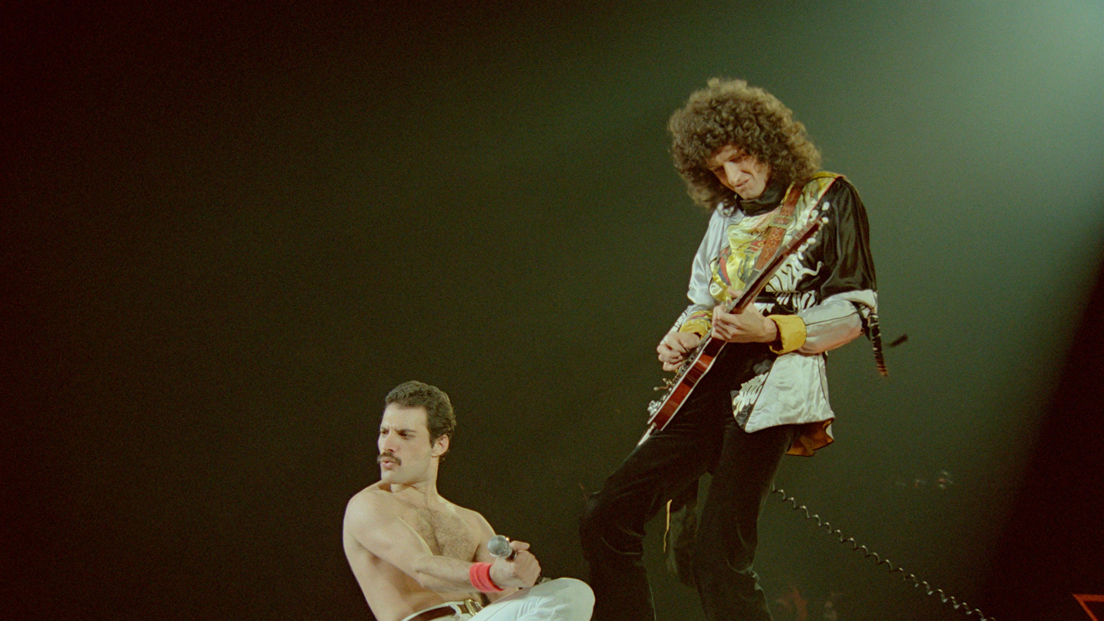 Freddy Mercury mit freiem Oberkörper und enger weißer Hose kniet, dahinter Brian May beim Gitarrensolo.