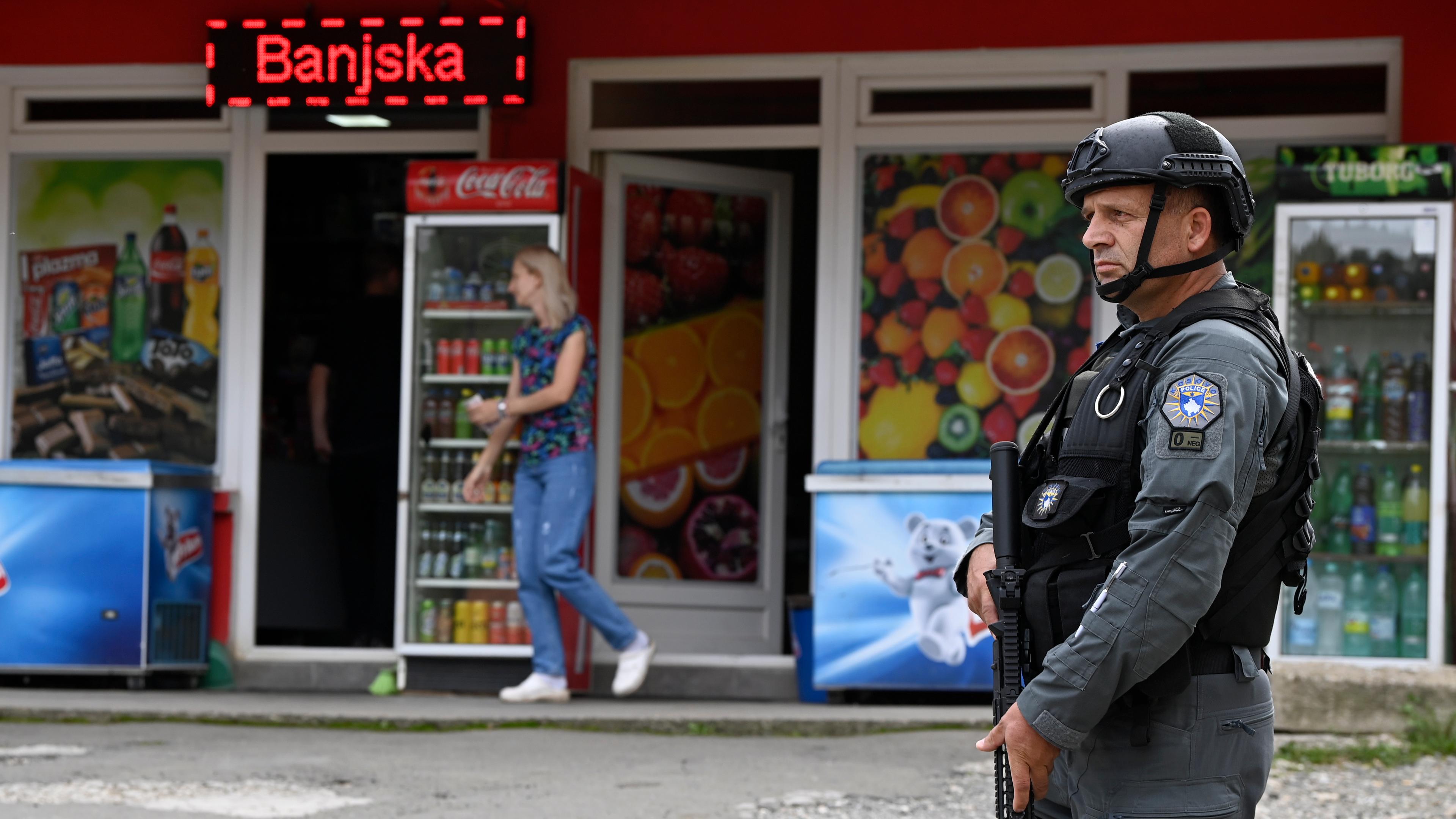 Ein kosovarischer Polizist in dunkler Uniform mit Gewehr, Helm und Schutzkleidung steht vor einem Supermarkt im Ort Banjska. Es leuchtet ein rotes Neon-Schild mit der Aufschrift "Banjska" im Hintergrund