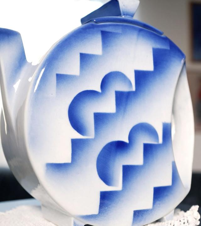 Das Bild zeigt eine dekorative Kanne, die auf einem runden Tisch steht. Die Kanne ist aus Keramik und hat eine weiße Grundfarbe, die mit blauen Mustern verziert ist. Die Muster bestehen aus geometrischen Formen, die an Zickzacklinien und Mondformen erinnern. Die Kanne hat einen länglichen Ausguss und einen kleinen Deckel.   Im Hintergrund ist eine unscharfe, helle Wand zu sehen, die wahrscheinlich ein Fenster darstellt, da Licht hereinströmt. Auf der linken Seite des Bildes ist eine schwarze Couch sichtbar, die mit einem weißen, filigranen Tuch gedeckt ist. An der Wand im Hintergrund hängen einige Rahmen mit Bildern, die jedoch nicht gut zu erkennen sind.   Das Bild vermittelt einen Eindruck von ebenso ästhetischen als auch kurvenreichen Linien und lädt zur Entdeckung der Sammlung ein.