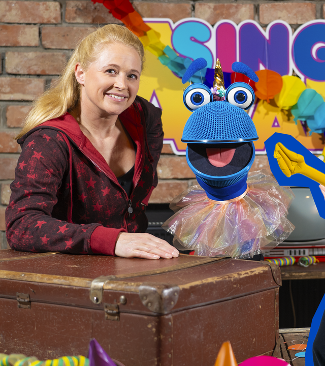 Singa und das SingDing schauen lachend hinter einem bunt dekorierten Tisch hervor. Das SingDing trägt ein buntes Tutu. Rechts von den beiden stehen Deniz & Ove, die mit aufblasbaren Gitarren posieren.