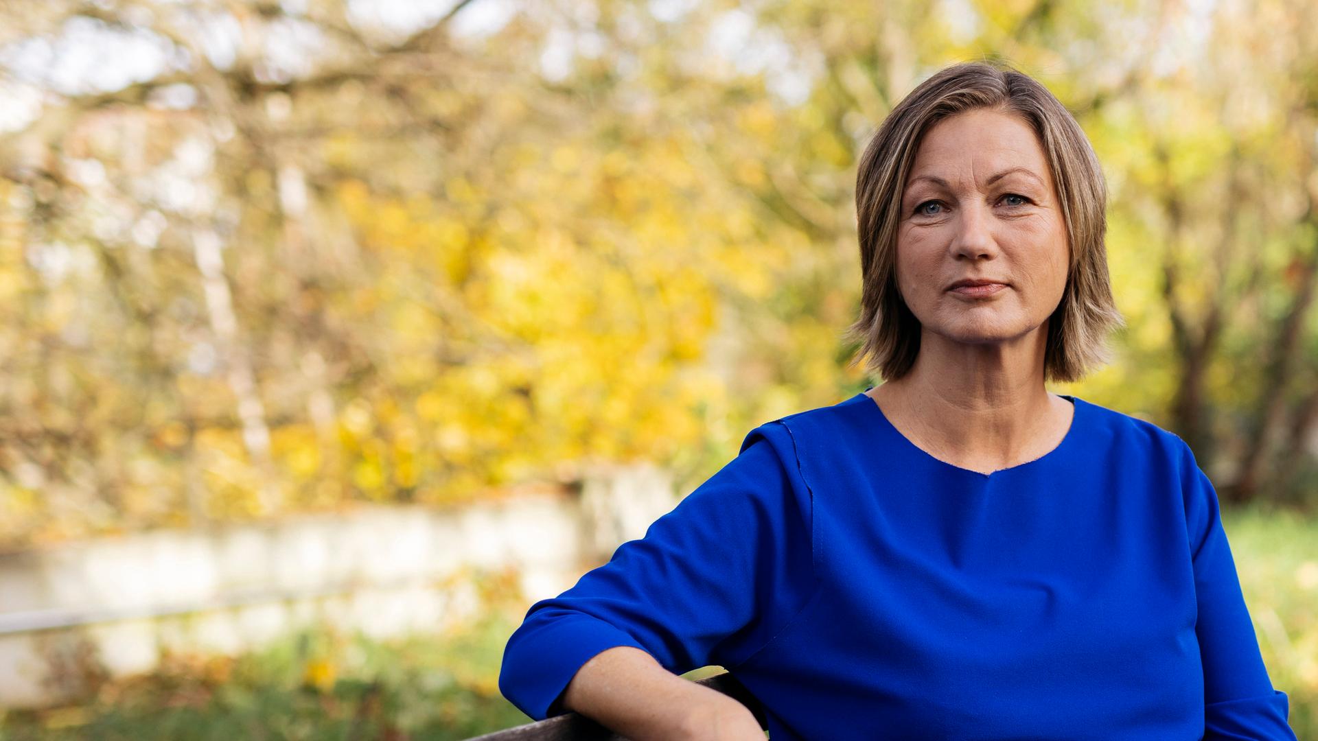 Silke Burmester sitzt auf einer Bank und schaut selbstbewusst in die Kamera. Im Hintergrund sind herbstlich anmutende Bäume zu sehen.