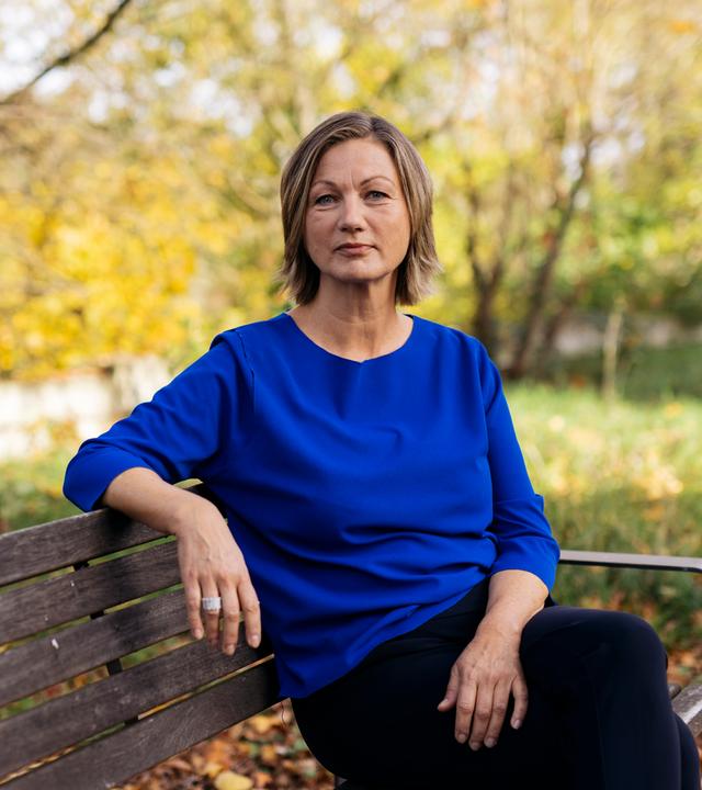 Silke Burmester sitzt auf einer Bank und schaut selbstbewusst in die Kamera. Im Hintergrund sind herbstlich anmutende Bäume zu sehen.