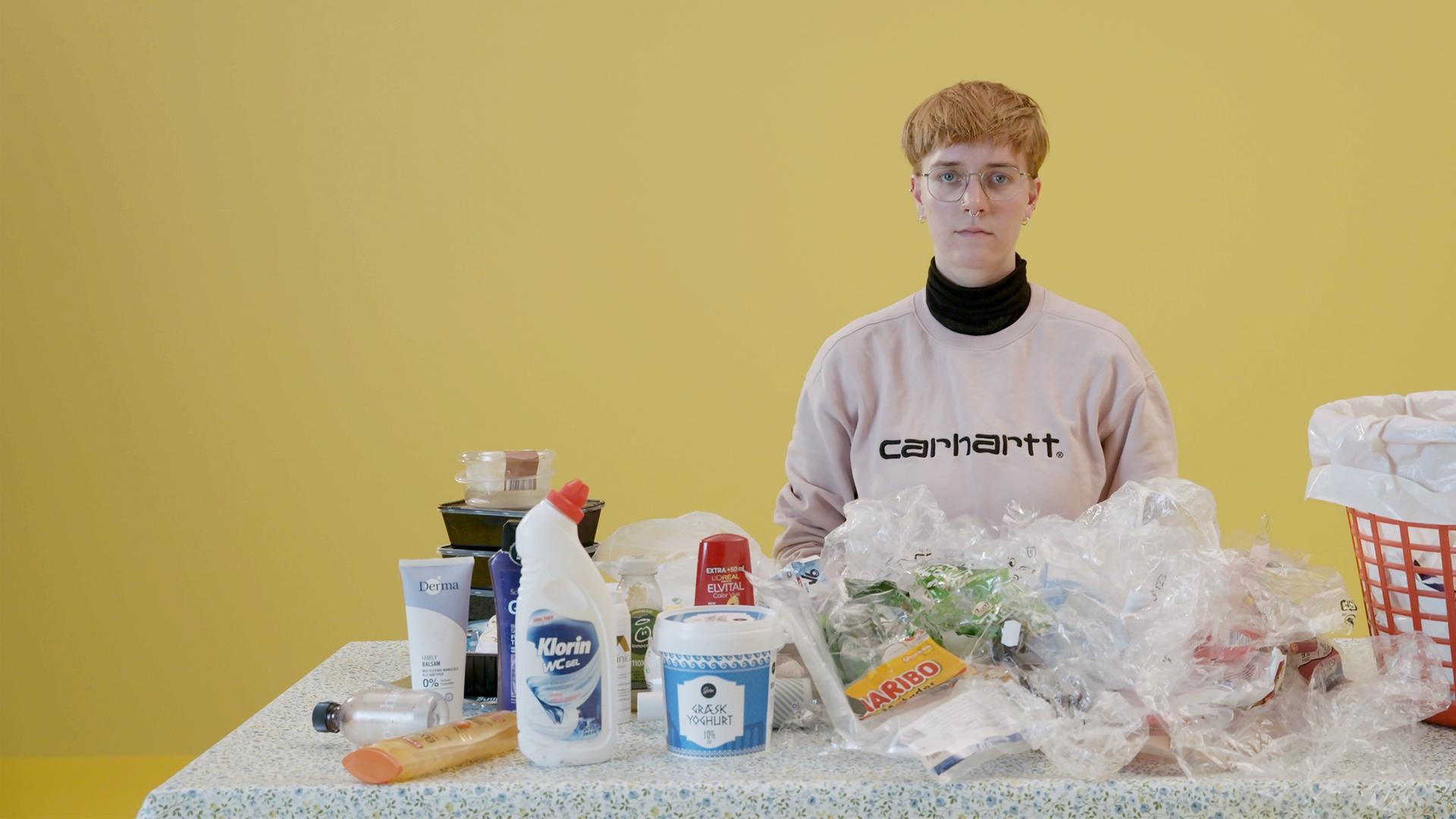 Eine junge Frau sitzt an einem Tisch, auf dem sich verschiedene Verpackungen aus Plastik befinden. Auf ihrer linken Seite steht ein großer Mülleimer.