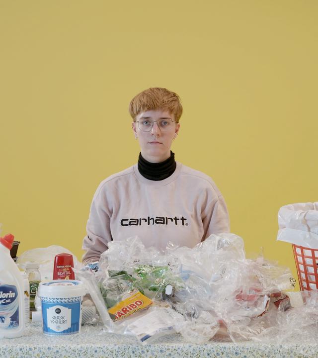 Eine junge Frau sitzt an einem Tisch, auf dem sich verschiedene Verpackungen aus Plastik befinden. Auf ihrer linken Seite steht ein großer Mülleimer.