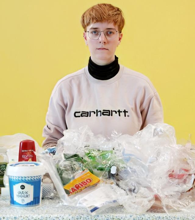Eine junge Frau sitzt an einem Tisch, auf dem sich verschiedene Verpackungen aus Plastik befinden. Auf ihrer linken Seite steht ein großer Mülleimer.