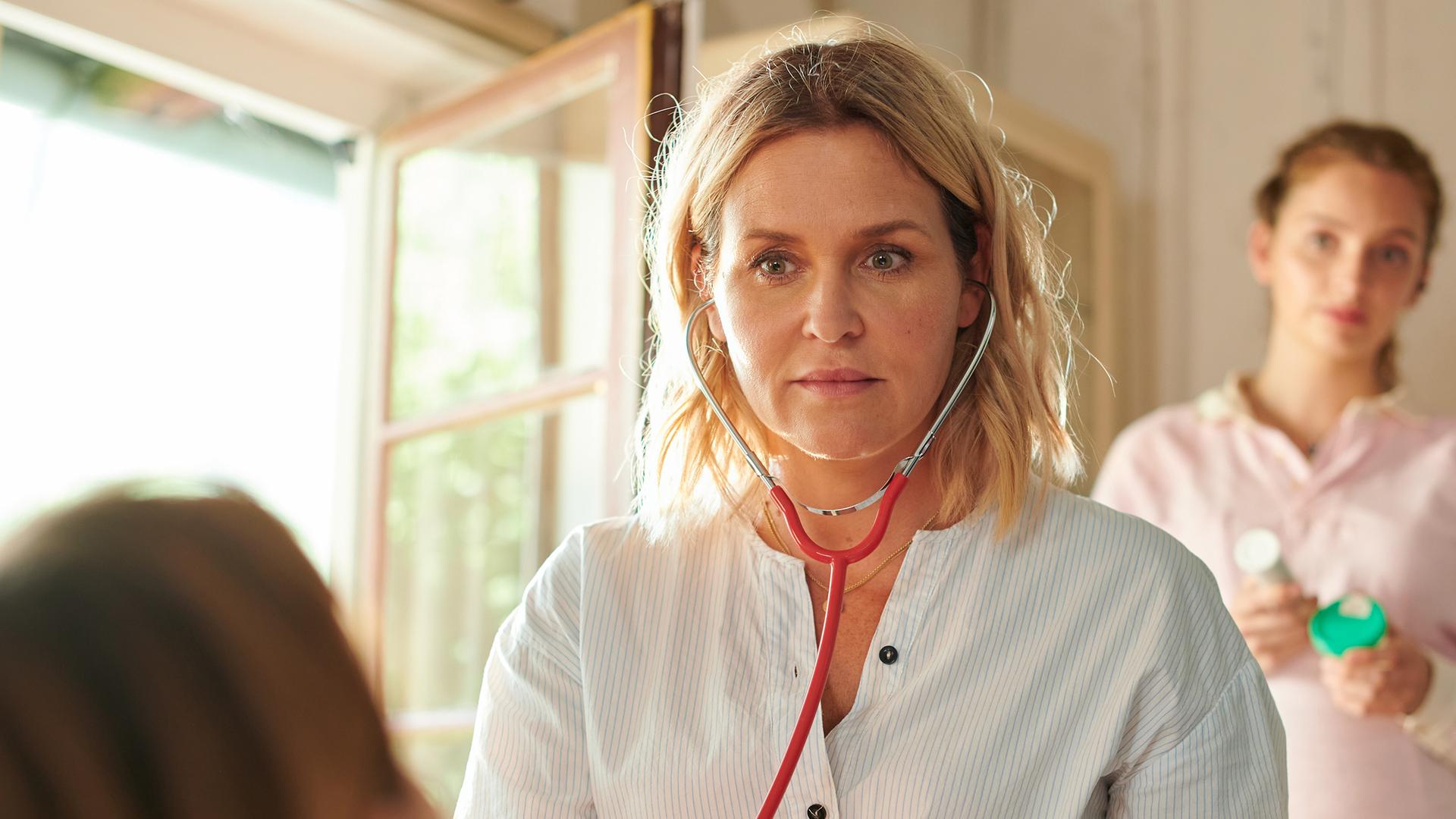 "Lena Lorenz - Krank vor Sorge": Auf einer Untersuchungsliege liegt Tati Meisinger (Anouk Elias) und blickt zur Decke. Lena Lorenz (Judith Hoersch) hat neben ihr Platz genommen und schaut sie besorgt an. Praktikantin Celine (Paulina Rümmelein) steht neben dem Kopfteil der Liege und beobachtet interessiert das Geschehen.