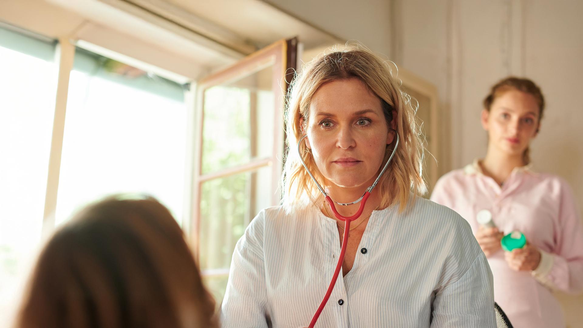 "Lena Lorenz - Krank vor Sorge": Auf einer Untersuchungsliege liegt Tati Meisinger (Anouk Elias) und blickt zur Decke. Lena Lorenz (Judith Hoersch) hat neben ihr Platz genommen und schaut sie besorgt an. Praktikantin Celine (Paulina Rümmelein) steht neben dem Kopfteil der Liege und beobachtet interessiert das Geschehen.