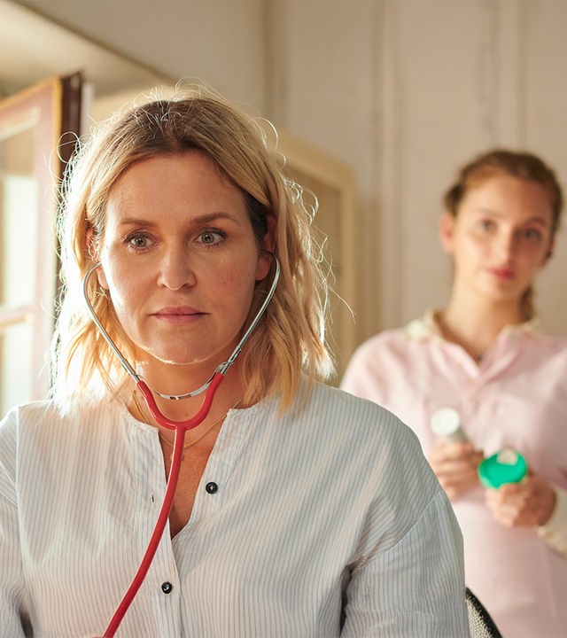 "Lena Lorenz - Krank vor Sorge": Auf einer Untersuchungsliege liegt Tati Meisinger (Anouk Elias) und blickt zur Decke. Lena Lorenz (Judith Hoersch) hat neben ihr Platz genommen und schaut sie besorgt an. Praktikantin Celine (Paulina Rümmelein) steht neben dem Kopfteil der Liege und beobachtet interessiert das Geschehen.