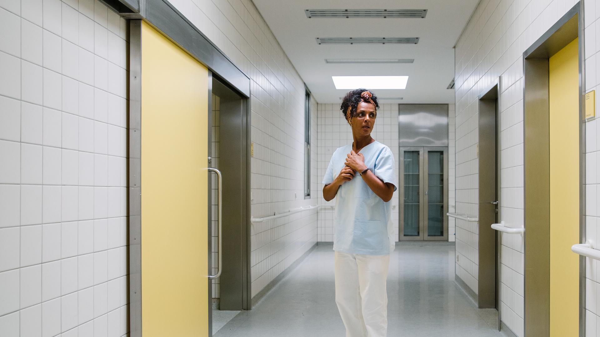 I don't work here: Lemlem (Dennenesch Zoudé) steht in weiß-hellblauer Pflegerinnenkleidung in der Mitte eines langen Krankenhausflures, an dessen Seiten gelbe Türen in andere Räume führen. Ihr Hände hält sie vor den Brustkorb und schaut zur Tür auf ihrer linken Seite.