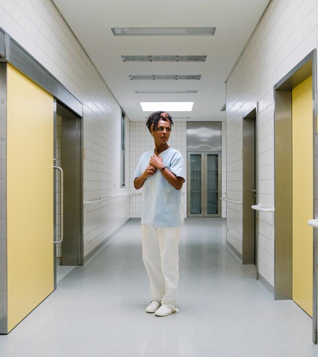 I don't work here: Lemlem (Dennenesch Zoudé) steht in weiß-hellblauer Pflegerinnenkleidung in der Mitte eines langen Krankenhausflures, an dessen Seiten gelbe Türen in andere Räume führen. Ihr Hände hält sie vor den Brustkorb und schaut zur Tür auf ihrer linken Seite.