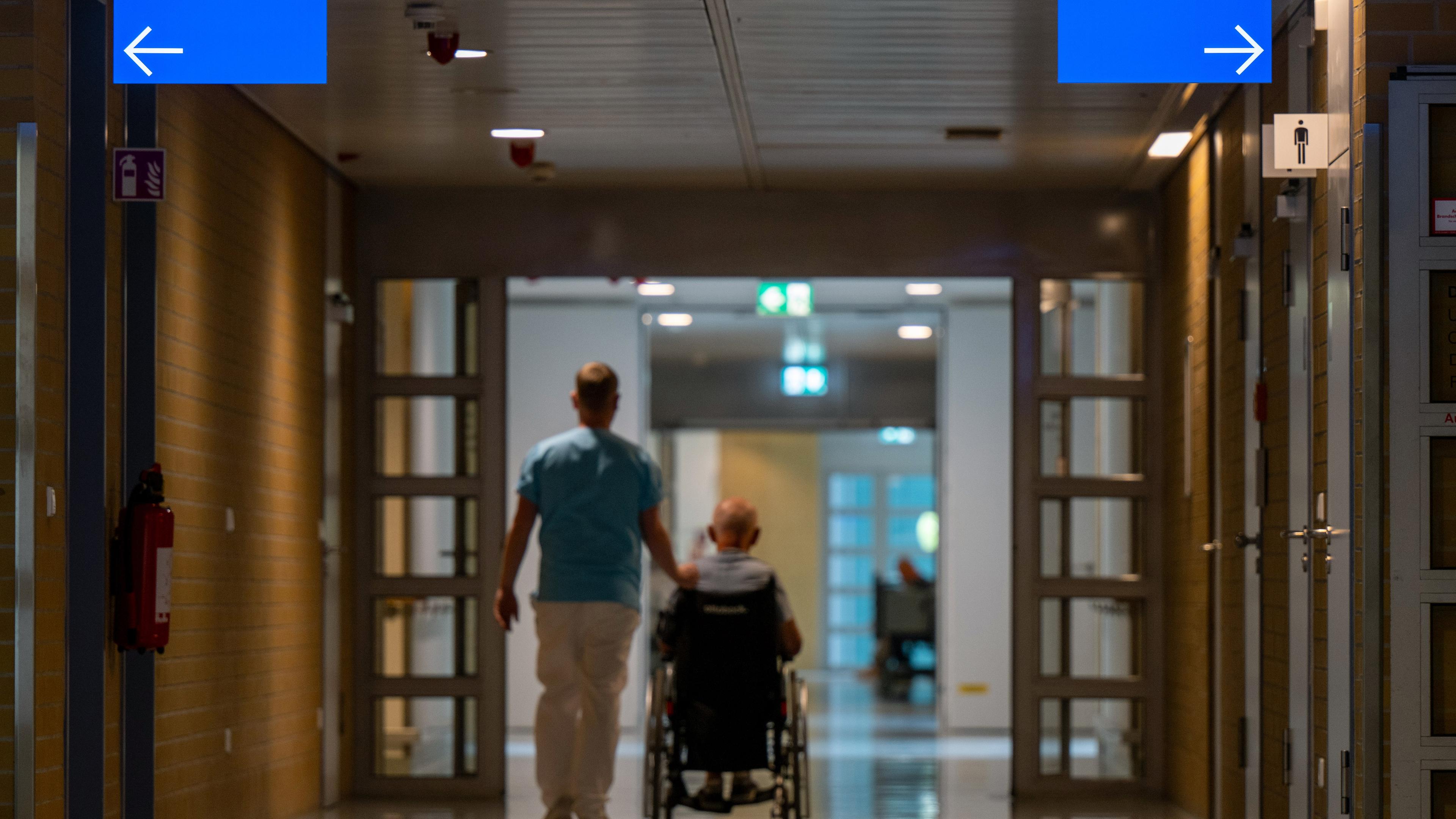 Flur in einem krankenhaus, von hinten sind ein Pfleger und ein Patient im Rollstuhl zu sehen.