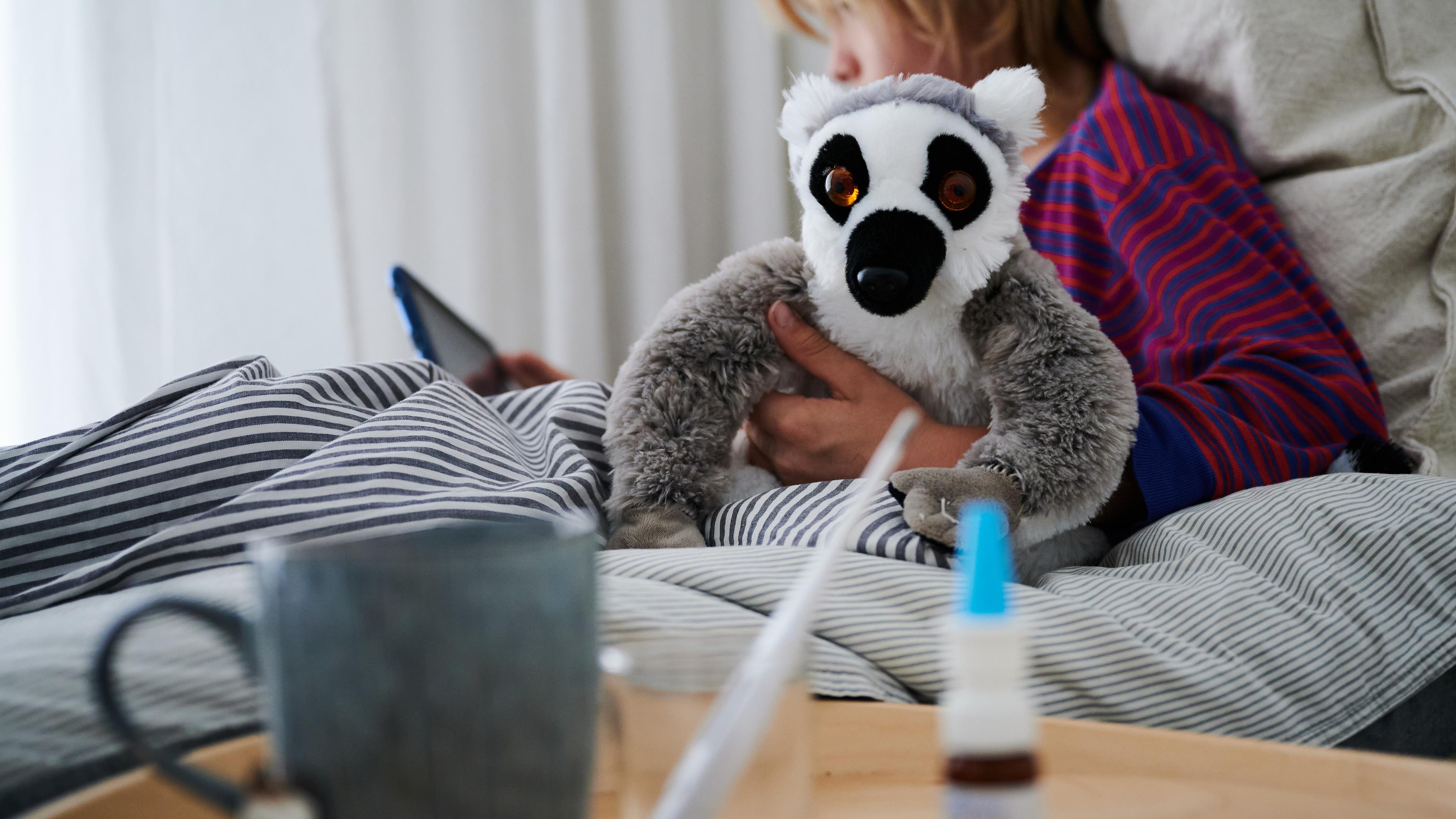 Ein Fieberthermometer, Nasenspray und eine Tasse Tee stehen vor einem Bett, in dem ein Kind mit Kuscheltier sitzt und auf ein Tablet schaut.