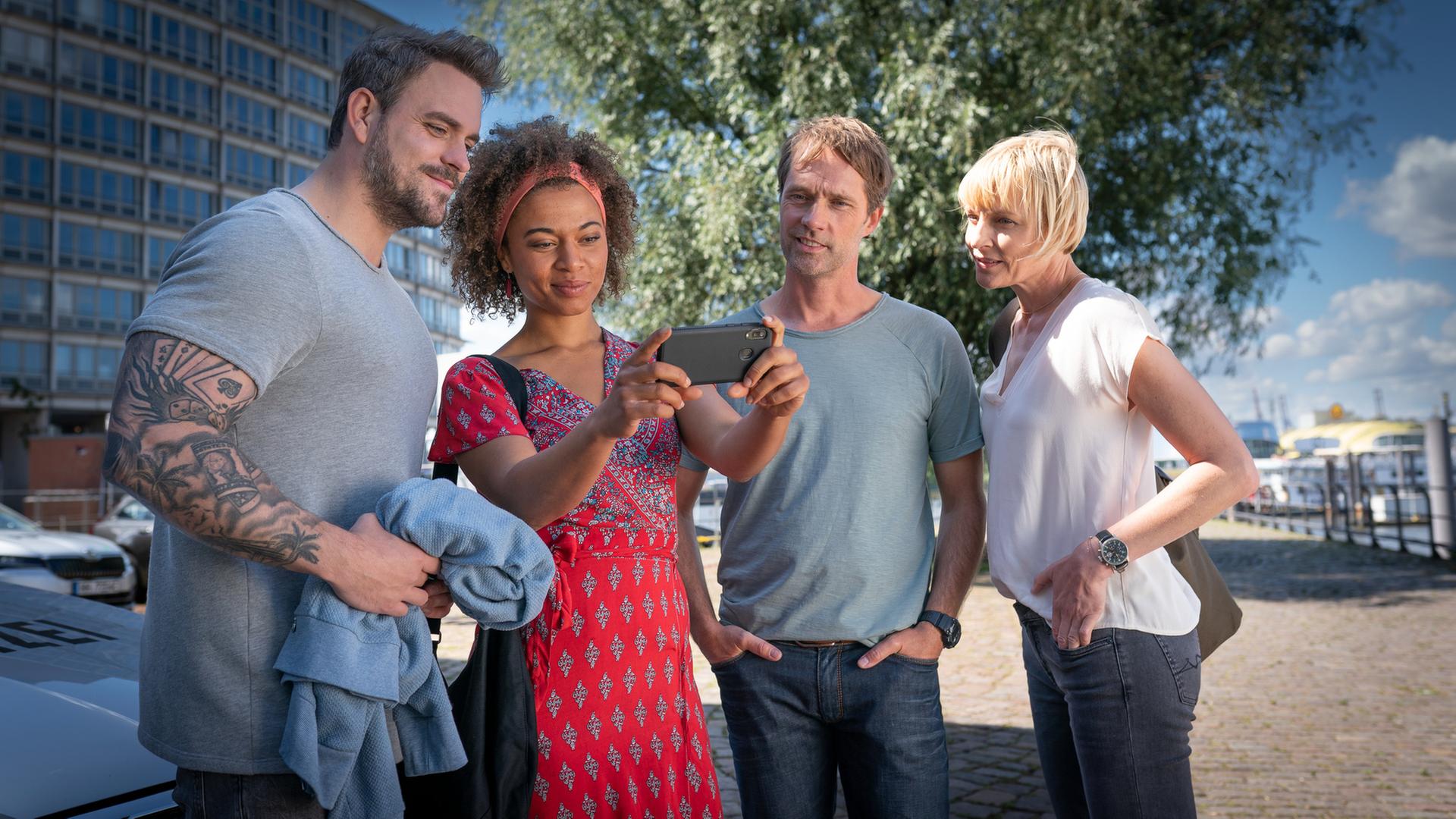 "Notruf Hafenkante - Krankhaft schön": Kris Freiberg (Marc Barthel), Daisy Petersen (Aysha Joy Samuel), Mattes Seeler (Matthias Schloo) und Melanie Hansen (Sanna Englund) stehen in Zivilkleidung im Freien nebeneinander. Daisy hält ein Handy vor sich, auf dessen Display alle schauen und lächeln.