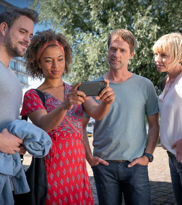"Notruf Hafenkante - Krankhaft schön": Kris Freiberg (Marc Barthel), Daisy Petersen (Aysha Joy Samuel), Mattes Seeler (Matthias Schloo) und Melanie Hansen (Sanna Englund) stehen in Zivilkleidung im Freien nebeneinander. Daisy hält ein Handy vor sich, auf dessen Display alle schauen und lächeln.