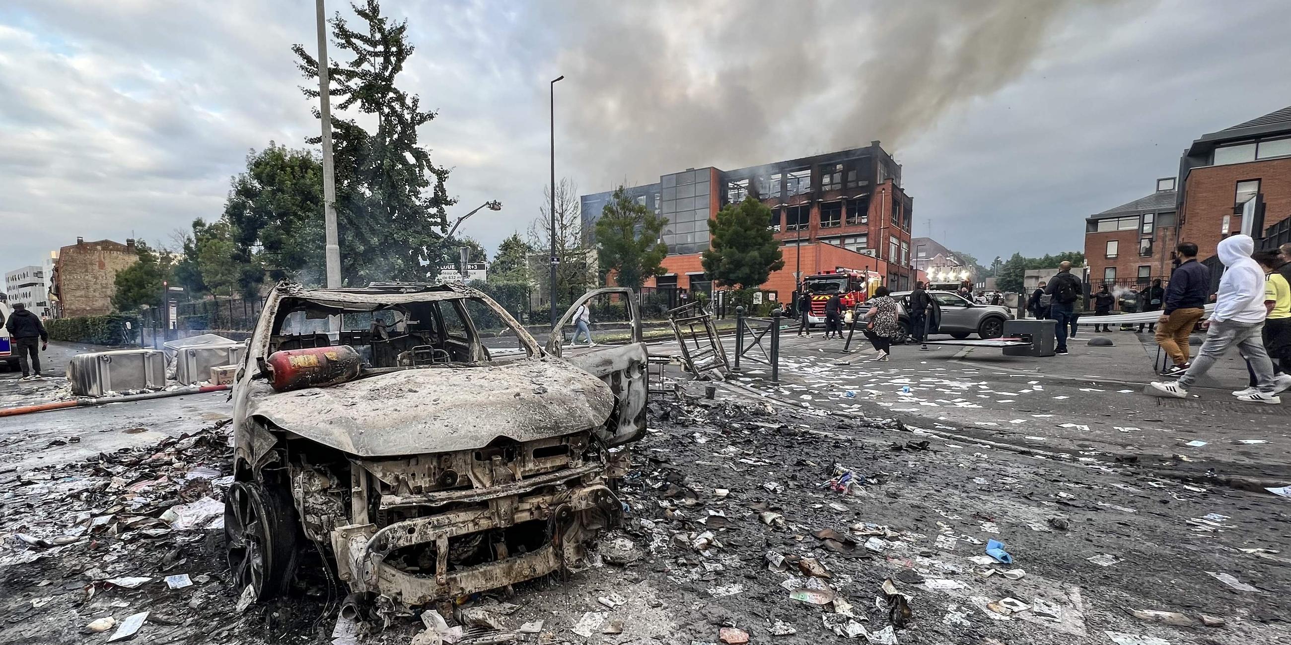 Ein ausgebranntes Fahrzeug steht auf einer Straße vor dem durch den Brand beschädigten Gebäude der Tessi-Gruppe im Stadtteil Alma von Roubaix, Frankreich