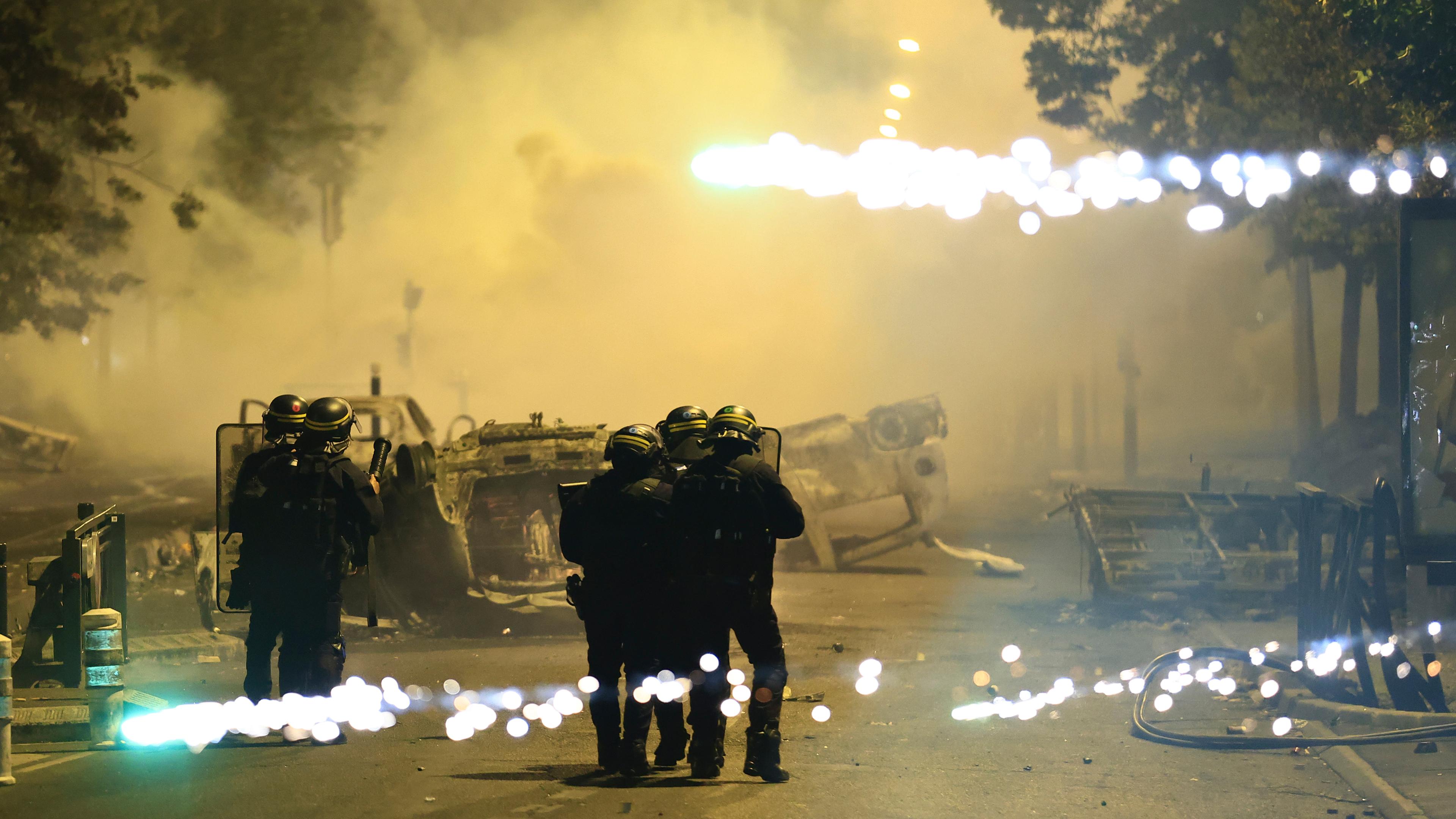 Beamte der Bereitschaftspolizei patrouillieren bei Unruhen auf einer Straße, während Feuerwerkskörper explodieren. 