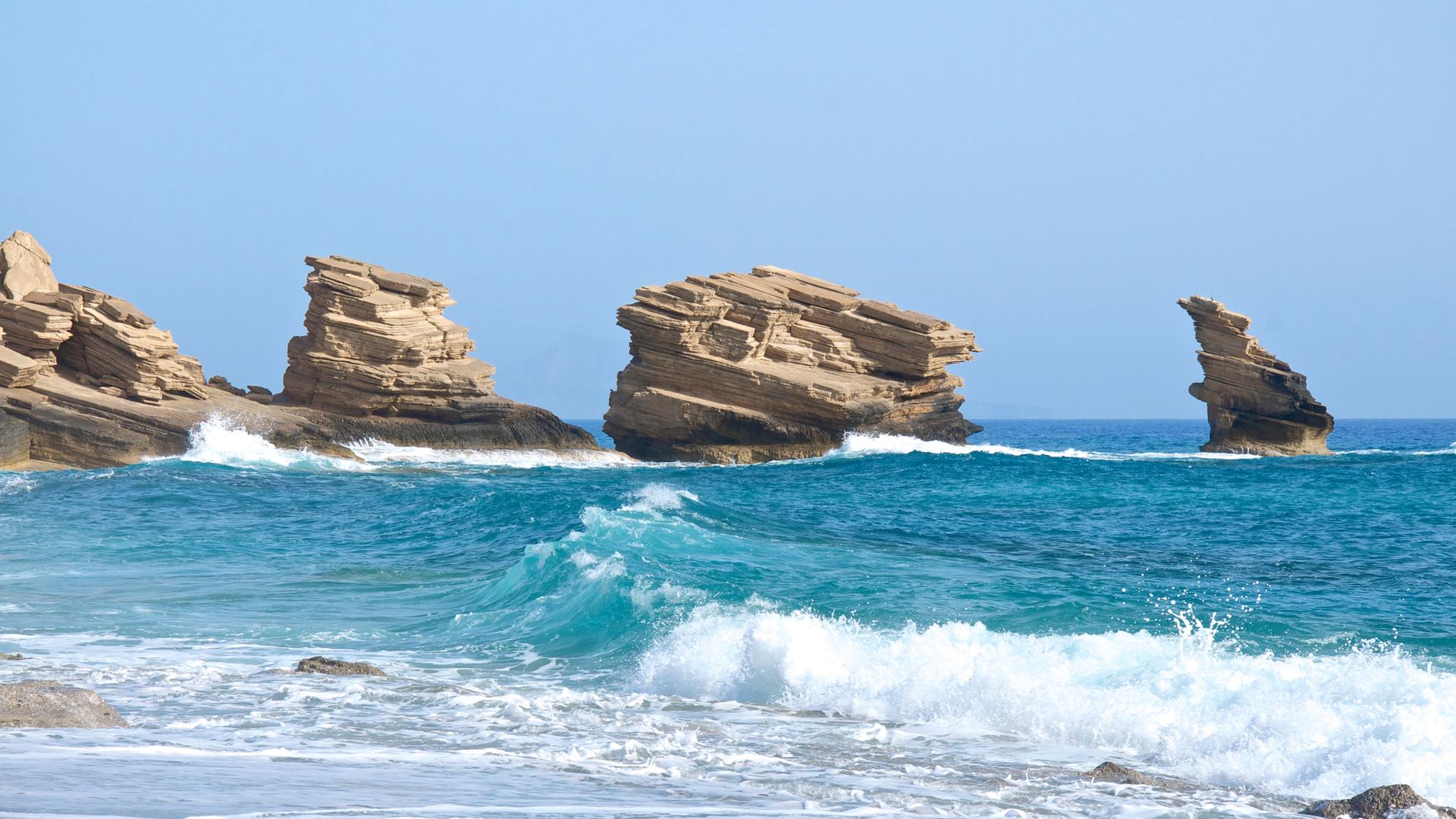 Wolkenloser blauer Himmel und ein Blick auf die Brandung am Strand von Triopetra auf Kreta. Im Hintergund stehen vier Felsen.