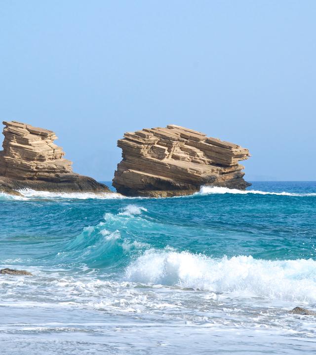 Wolkenloser blauer Himmel und ein Blick auf die Brandung am Strand von Triopetra auf Kreta. Im Hintergund stehen vier Felsen.
